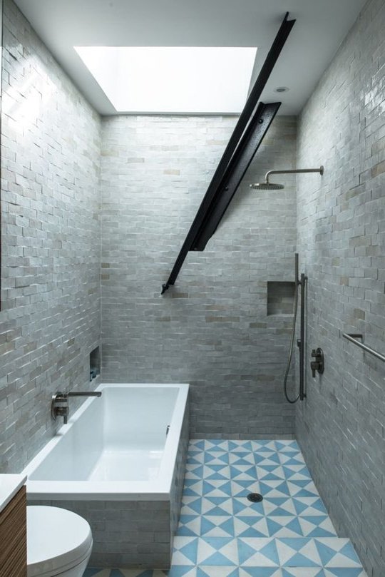 baño moderno pequeño con paredes de piedra gris, suelo con diseños triangulares celeste y blanco, bañera blanca