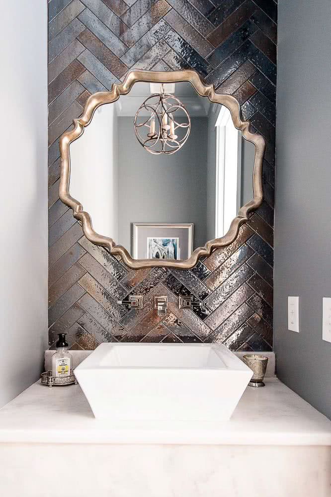 lavabo y encimera de cuarzo blanco, pared posterior con azulejos en espiga en acabado metálico en colores plateados y bronce. Espejo decorativo con marco color oro