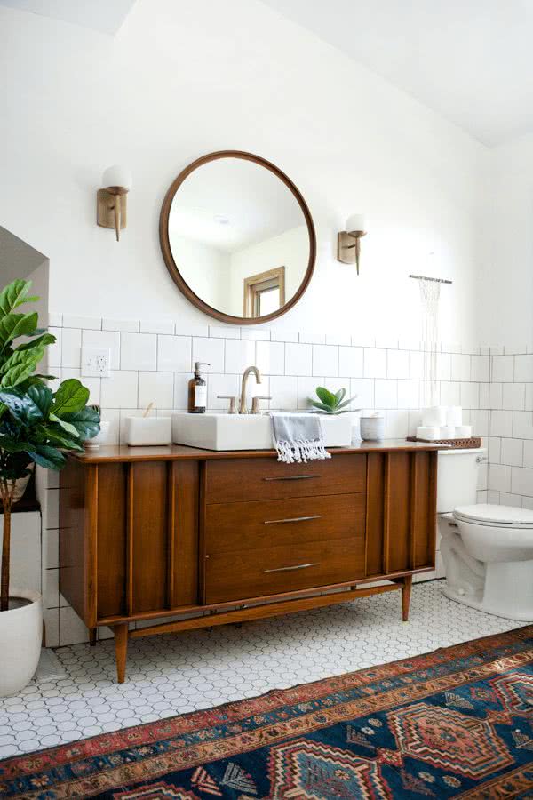armario bajo de mediados de siglo en madera, lavabo rectangular, espejo redondo con marco dorado, paredes y suelo blanco