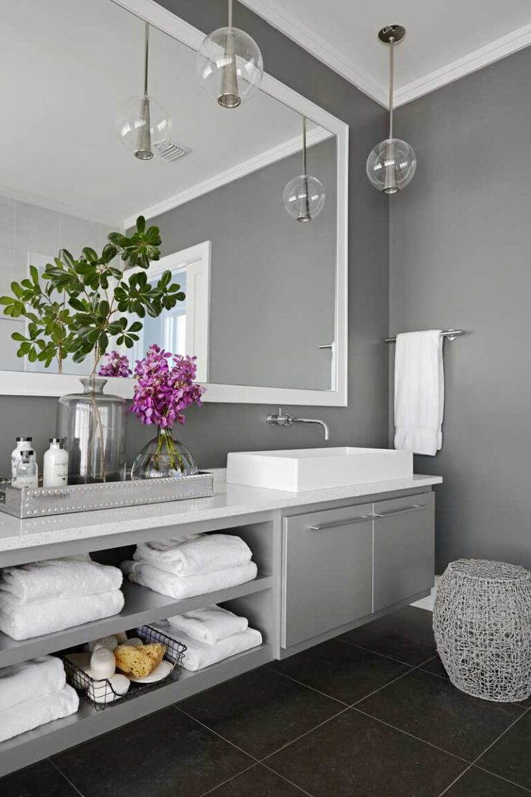 baño con paredes grises, espejo con marco blanco, muebles bajos en color gris con estantes abiertos